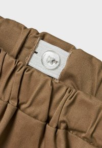 Brun elastisk midjeband med texturerad tyg, med en vit etikett med en knapp. Volangdetaljer förbättrar designen.