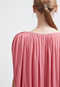 Różowa, miękka bluzka z gumką przy dekolcie i luźnym kroju. Widok z tyłu ukazuje gładką teksturę i subtelny efekt drapowania.