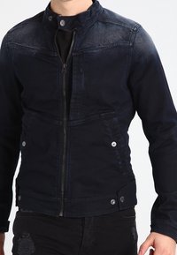 Man som bär en mörkblå zip-up jacka med knäppt krage och fickor, ihop med slitna svarta jeans.