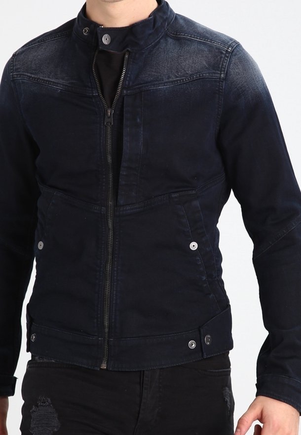 Man som bär en mörkblå zip-up jacka med knäppt krage och fickor, ihop med slitna svarta jeans.