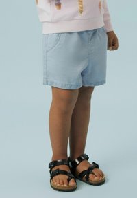 Shorts de algodón azul claro con un ajuste relajado, que cuentan con bolsillos laterales y un dobladillo sencillo. Combinados con sandalias negras con hebilla sobre un fondo claro.