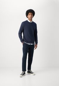 Marineblauwe sweater met ronde hals, lange mouwen, gecombineerd met donkerblauwe broek en zwarte hoge sneakers met witte zolen en veters.