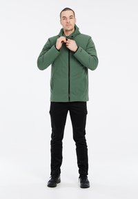 Chaqueta verde con capucha y cremallera frontal, hecha de un material suave, combinada con pantalones ajustados negros y calzado oscuro.