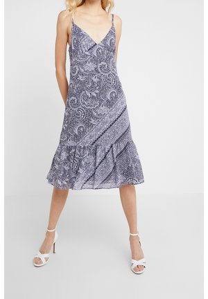 Frau trägt ein ärmelloses Midi-Kleid mit V-Ausschnitt und schwarz-weißen Paisley- und Blumenmustern, kombiniert mit weißen Sandalen mit hohen Absätzen.