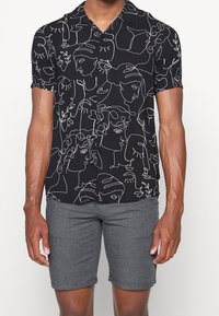 Uomo con una camicia nera a maniche corte con bottoni, decorata con un'illustrazione astratta di un volto in bianco, e pantaloni corti grigi, su uno sfondo bianco.