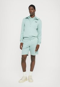 Mintgrønn treningsdrakt laget av mykt stoff, med en glidelåsjakke og shorts. Inkluderer en logo på brystet og teksturerte detaljer på skoene.