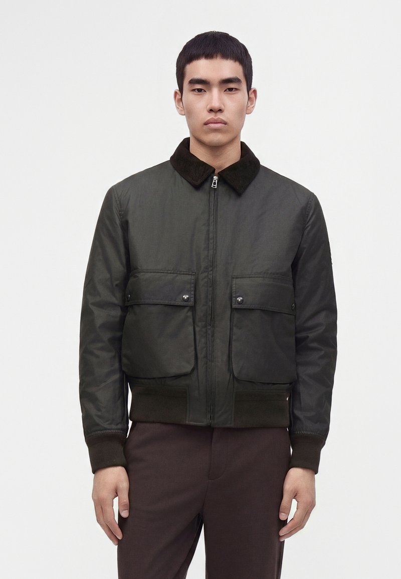 Jeune homme aux cheveux courts et foncés, portant une veste zippée vert foncé avec des poches à rabat et un pantalon marron foncé, face à l’avant.