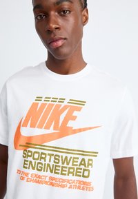 Bijela pamučna majica s pomožnim narandžastim i zelenim Nike logom i natpisom, okrugli izrez, kratki rukavi i opušteni kroj.
