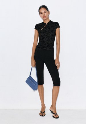 Femme portant un haut noir à manches courtes à motif, des leggings noirs longueur genou, des sandales à talons noires, tenant un sac à main bleu à ses côtés.