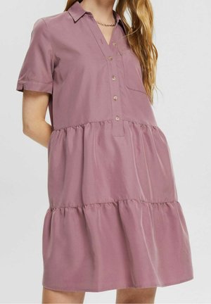 Kobieta w krótkim, mauve sukience na guziki, z kołnierzykiem i przednią kieszenią, stojąca na tle jasnego, jednolitego tła.