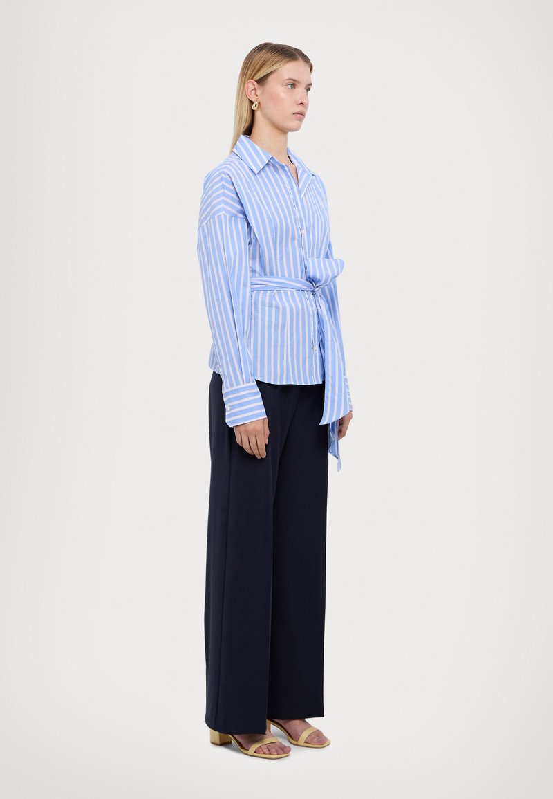 Lichtblauw en wit gestreept shirt met een knoop in de taille, gedragen met marineblauwe wijde broek en gele hoge hakken sandalen.