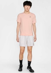 Camiseta de manga corta rosa claro con un pequeño logo, combinada con pantalones cortos deportivos gris claro que cuentan con bolsillos laterales. Zapatillas deportivas y calcetines negros.