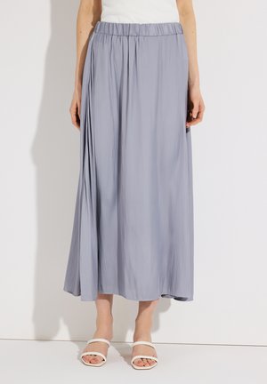 Frau trägt einen hellblauen, plissierten Maxirock, ein weißes ärmelloses Oberteil und weiße Sandalen mit geöffneten Zehen und zwei Riemen.