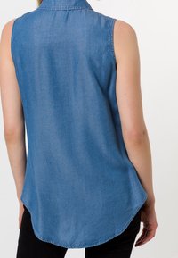 Sleeveless denim shirt in light blue with a collared neckline, curved hem, and a smooth texture. Visible stitching details on the seams.