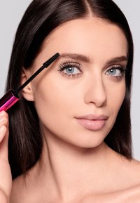 Spazzola per mascara nera tenuta sopra sopracciglia definite. Tubo di mascara rosa lucido riflette la luce. Modella con pelle chiara e capelli lisci e scuri.