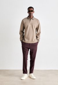 Beige long-sleeve polo with a collar and logo; paired with dark burgundy trousers and white sneakers, showcasing a relaxed fit.