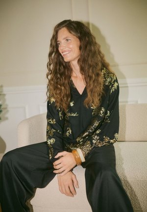 Une femme souriante aux longs cheveux bruns ondulés est assise sur un canapé beige. Elle porte une tenue noire ornée de motifs floraux dorés et des bracelets.
