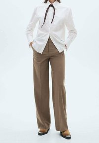 Chemise blanche boutonnée avec deux poches poitrine, pantalon large marron, et chaussures à talons noires avec un accent métallique. Écharpe marron nouée au col.