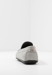 Chaussure à enfiler en daim gris avec un dos arrondi, des coutures décoratives et une petite étiquette rouge "HUGO" au talon. Semelle plate en caoutchouc noir.