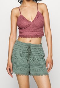 Kvinna i spets crop top i askros och matchande spets shorts i salviagrön, stående framför en ljus, enfärgad bakgrund.