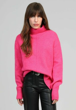 Femme aux longs cheveux bruns portant un pull à col roulé rose vif et un pantalon en cuir noir sur un fond gris uni.