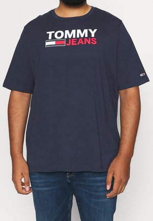 Camiseta de manga corta azul oscuro con el logo "TOMMY JEANS" en blanco y rojo en el pecho, llevada con jeans azules por una persona de tono de piel medio.