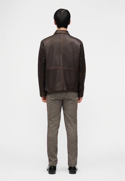 Άνδρας με σκούρο καφέ δερμάτινο μπουφάν, γκρι υφασμάτινο παντελόνι με υφή και μαύρα παπούτσια, στέκεται με την πλάτη προς το μέρος μας μπροστά σε απλό λευκό φόντο.