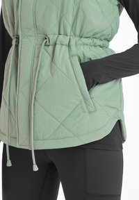 Quilted light green vest with adjustable drawstring waist, side pockets, and contrast black long sleeve underneath.