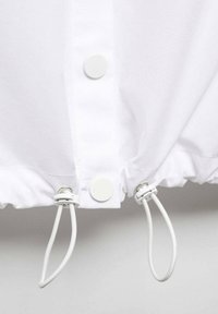 Giubbotto antivento bianco con orlo aderente regolabile mediante coulisse elastiche, chiusura con bottoni a pressione e tessuto dalla texture liscia.