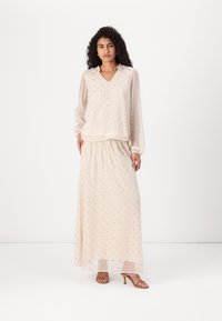 Blusa leggera color beige chiaro, trasparente, con scollo a V, maniche lunghe e delicati pois ricamati, abbinata a una gonna lunga e coordinata.