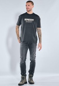 Man with tattoos wearing black "REPRESENT AUTHENCITY" t-shirt, dark jeans, and gray boots, standing against a plain light background.