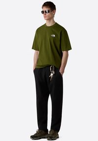 Camiseta de algodón verde oliva con un pequeño logo blanco, combinada con pantalones negros de corte holgado y zapatillas gris-marrón. Accesorio incluye llavero.