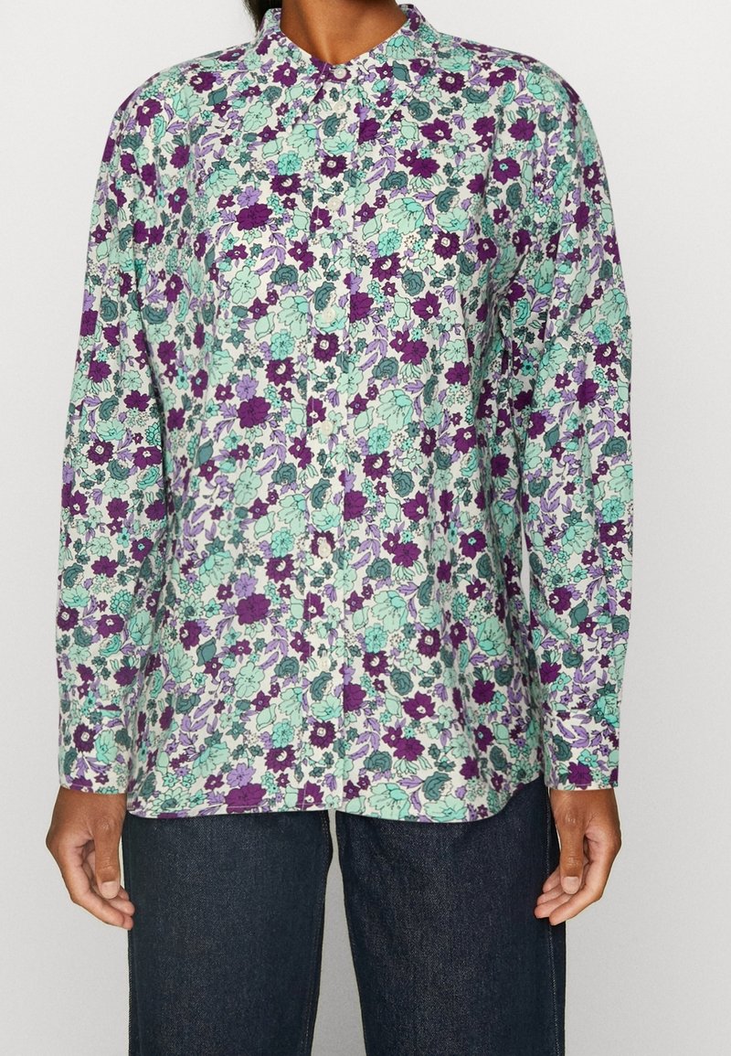 Chemise à manches longues boutonnée avec un motif floral dense en violet, vert et lavande, portée avec un jean bleu foncé.