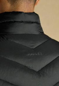 Čierna prešívaná bunda s vysokým golierom, so vzorom v tvare šípok a jemným embosovaným logom "JOULES" na hornej časti chrbta. Hladká textúra.