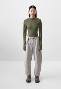 Haut vert à manches longues avec des manches texturées, associé à un pantalon léger gris clair et ample avec une ceinture bleue. Des chaussures de randonnée marron complètent le look.