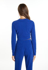Blå crop top med lange ærmer, lavet af glitrende stof. Har en rund halsudskæring og en tætsiddende silhuet, parret med matchende bukser.