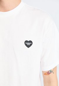 White cotton t-shirt with short sleeves featuring a black heart-shaped patch with "Carhartt WIP" text, positioned on the left chest area.