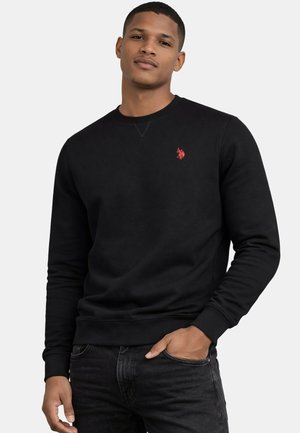 Junger Mann trägt einen schwarzen Langarm-Sweatshirt mit einem kleinen roten Emblem auf der Brust, die Hände entspannt, vor einfachem Hintergrund.