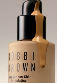 Bobbi Brown Weightless Skin Foundation i en rund, beige flaske med en sort pumpe, med en strømmende mengde produkt i en brun nyanse.