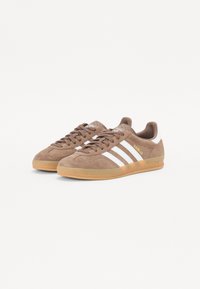 Hnědé semišové tenisky s bílým designem tří pruhů a žlutým textem "Gazelle". Guma z kaučuku, kulatá špička a klasické šněrování vpředu.