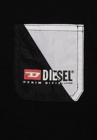 Černá látka s kapsou na levé hrudi, která má diagonální bílo-černý design, označená logem "DIESEL DENIM DIVISION".