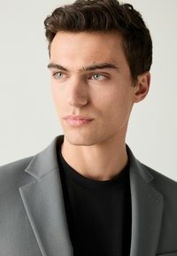 Harmaa bleiseri, jossa on sileä pinta, notched-lapel ja yksittäinen nappi, mustan pyöreäkauluksisen paidan päällä. Kuva otettu lähietäisyydeltä.