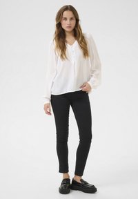 Bílá blůza s dlouhým rukávem, výstřihem do V a detaily knoflíků, spárovaná s černými skinny džínami a černými nazouvacími botami. Hladká struktura látky.