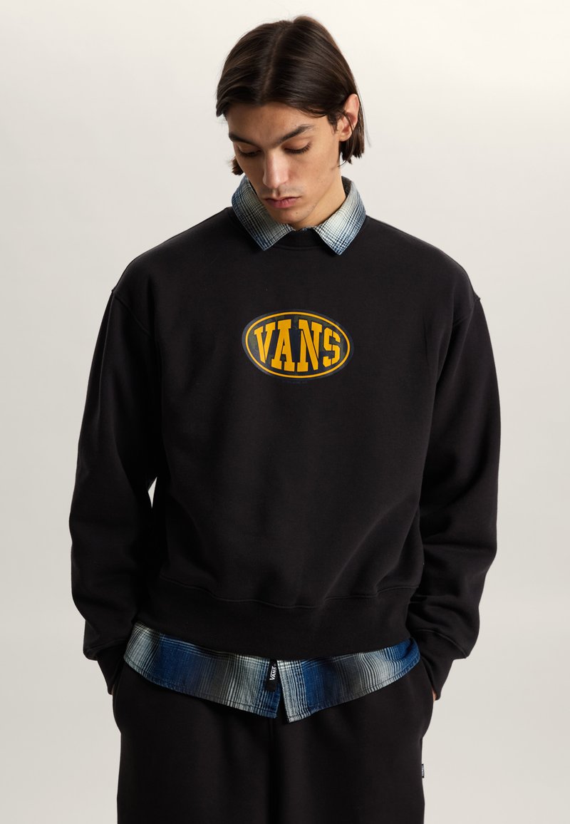 Young man wearing a black Vans sweatshirt over a blue plaid shirt, hands in pockets, looking down against a plain background.
