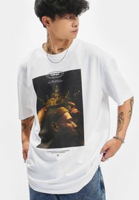 Ung mand med tatoveringer og smykker iført en hvid oversized T-shirt med et portræt af en kronet mand og tekst.