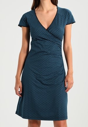 Woman wearing a short-sleeve, blue patterned wrap dress with a V-neckline, standing against a plain light background.