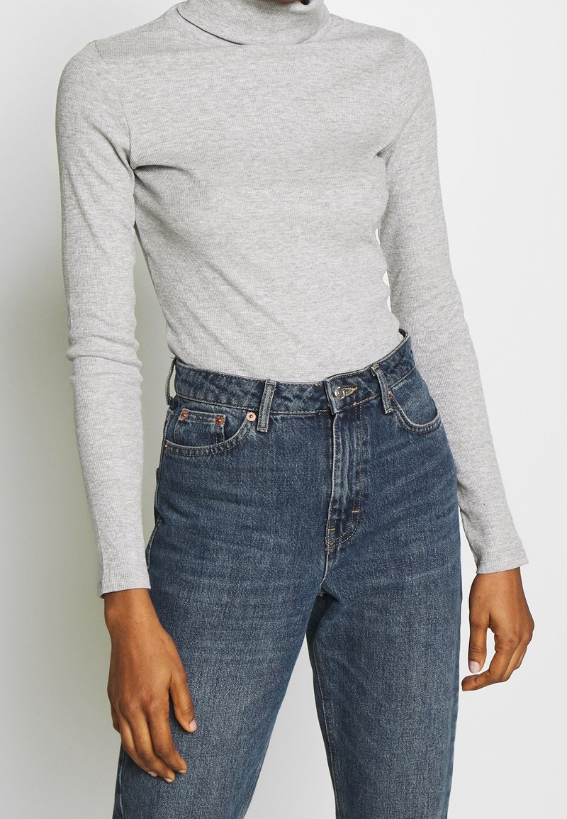 Personne portant un col roulé gris clair à manches longues rentrée dans un jean en denim bleu taille haute, les mains détendues le long du corps.