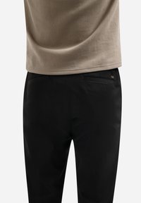 Achteraanzicht van een persoon in een beige shirt en zwarte broek met twee achterzakken tegen een witte achtergrond.