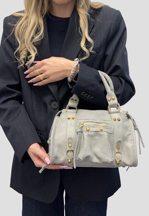 Donna con capelli biondi lunghi, indossa un blazer nero e tiene una borsa in suede grigio chiaro con borchie dorate e dettagli con cerniera.