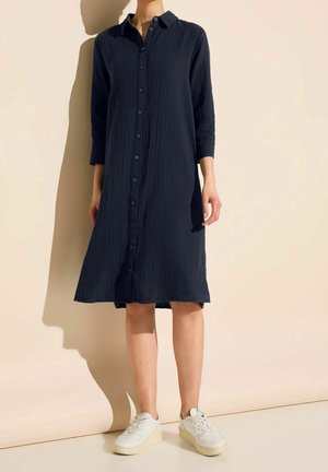 Femme portant une robe noire boutonnée, à manches longues, longueur genou, associée à des baskets blanches, debout contre un mur beige clair.